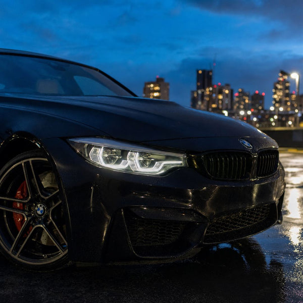 Phare LED d'une BMW M3 noire allumé de nuit, anneaux de feux de jour et logo BMW sur le capot