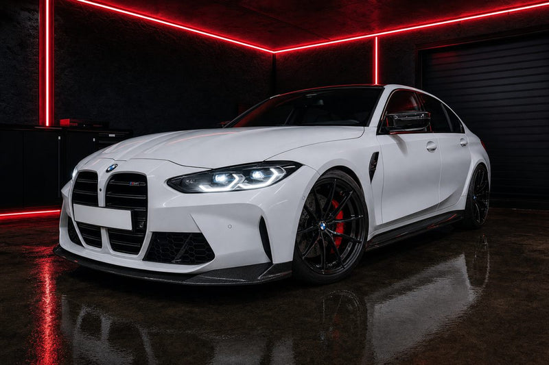 BMW M3 G80 blanche modifiée, lame avant et coques de rétroviseur en carbone, dans un garage éclairé par des LED rouges
